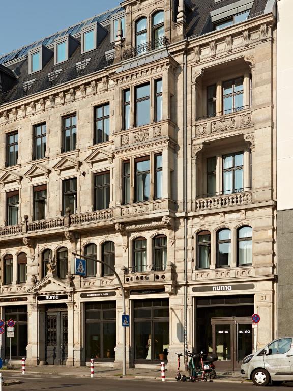 Boutique Hotels in Berlin Kreuzberg 17 A historic multi-story stone building with ornate architectural details and large windows; the ground floor features entrances to boutique hotels in Berlin Kreuzberg, with street signs, parked scooters, and bollards visible on the sidewalk.