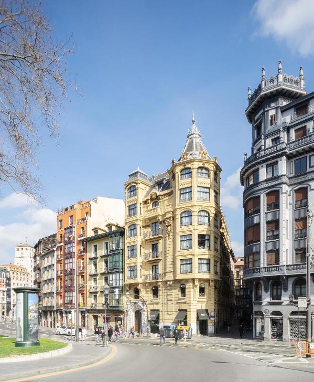 8 Unique Boutique Hotels in Bilbao Spain 9 A city street curves past ornate historic buildings showcasing varied architecture; people stroll wide sidewalks under a blue sky with scattered clouds, just steps from some of the finest boutique hotels in Bilbao.