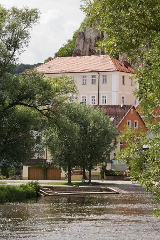 FAIRYTALE Castle Hotels in Bavaria 7 A river flows past trees and a large, light-colored building with a red-tiled roof—reminiscent of the charming castle hotels in Bavaria—nestled at the base of a rocky hill and partly hidden by lush greenery.