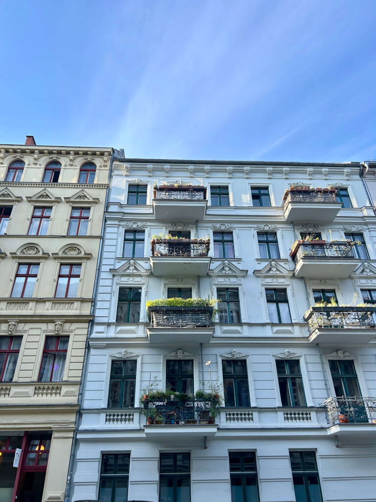 Boutique Hotels in Berlin Kreuzberg 2 Two adjacent European-style buildings, reminiscent of boutique hotels in Berlin Kreuzberg, feature ornate balconies. The left is beige; the right is white with green plants on balconies, all set against a clear blue sky.