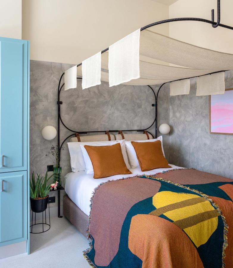 15 Best Boutique Hotels in London, UK 26 A cozy bedroom reminiscent of the best boutique hotels London offers, with a four-poster bed, brown and mustard bedding, white pillows, and canopy drapes. Gray walls, pastel artwork, a blue wardrobe, and a green potted plant complete the scene.