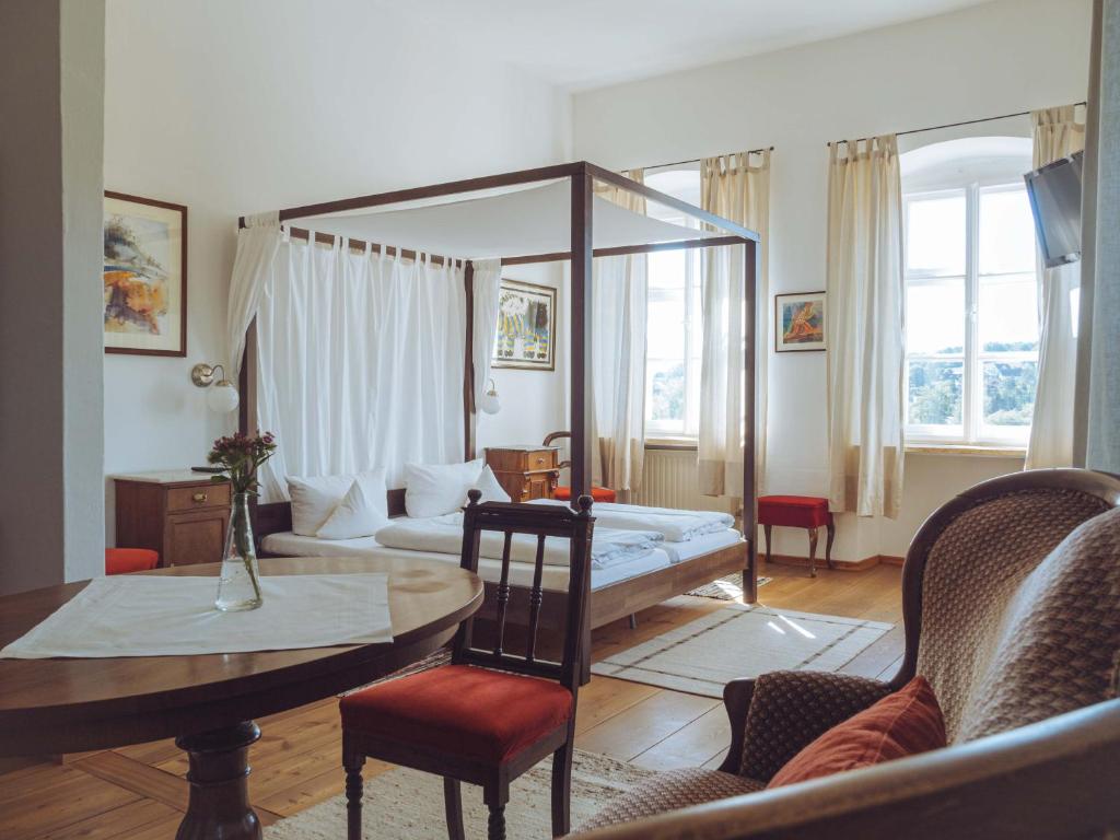 FAIRYTALE Castle Hotels in Bavaria 8 A bright, elegant bedroom inspired by castle hotels in Bavaria, featuring a canopy bed, wooden floor, tall windows with light curtains, classic wooden furniture, and a round table with a vase of flowers. Sunlight creates a cozy atmosphere.