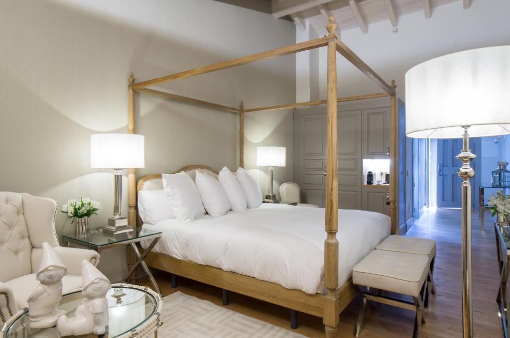 A stylish bedroom inspired by boutique hotels in Asturias, featuring a wooden four-poster bed, white bedding, modern lamps, benches at the foot, a side table with flowers, an armchair, and decorative rabbit figures on a glass table in soft neutral tones.
