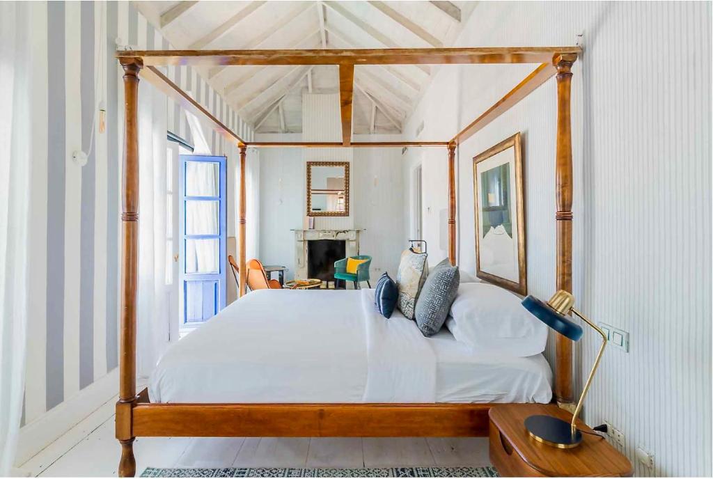 Adults-Only Hotels in Costa del Sol 17 A bright bedroom in the style of adult only hotels in Costa del Sol, featuring a wooden four-poster bed, white linens, striped walls, a blue door, a desk lamp, framed art, a mirror above the fireplace, and high beamed ceilings.