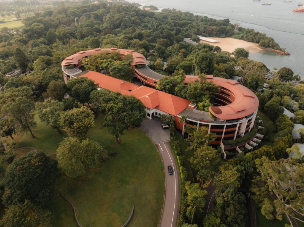 13 Most Unique Hotels in Singapore for an Incredible Holiday 52 Aerial view of a large, curved building with a red roof surrounded by lush greenery, near a body of water with anchored ships and a sandy beach in the background—one of the unique hotels in Singapore with scenic surroundings. A road leads to the entrance.