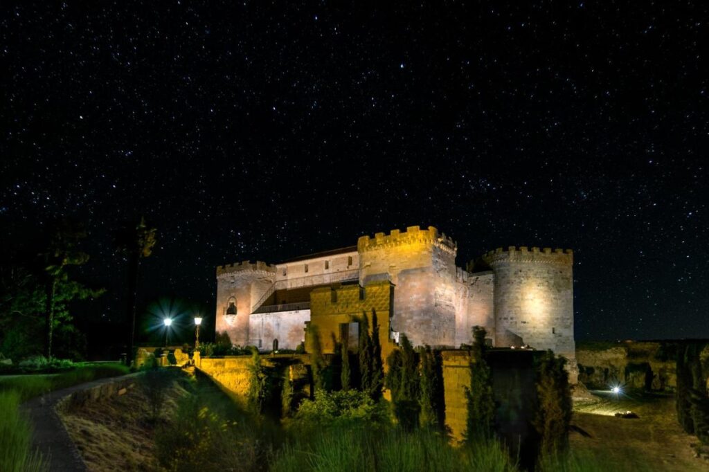 25 Weekend Trips from Madrid Max. 2h Away 29 A large stone castle illuminated at night, surrounded by trees and greenery, with a clear, star-filled sky in the background—an enchanting spot perfect for weekend trips from Madrid.