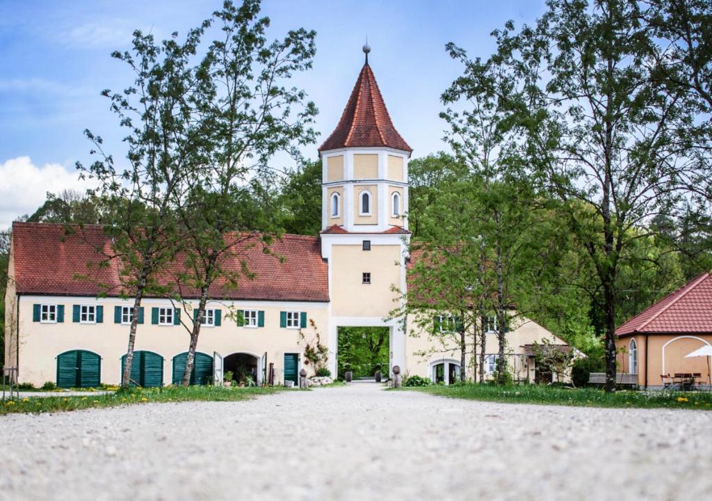 FAIRYTALE Castle Hotels in Bavaria 25 A path leads to a pale yellow building with a tall, pointed tower and red-tiled roofs, reminiscent of castle hotels in Bavaria, surrounded by trees and greenery under a blue sky.