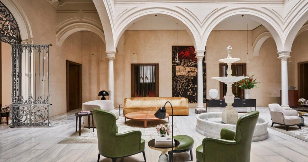 10 UNIQUE Hotels with Pool in Seville 15 Sophisticated lounge with green armchairs, a tan sofa, marble floors, and a central white fountain under arched ceilings. Ornate iron gates and modern decor capture the elegance of top hotels with pool in Seville.