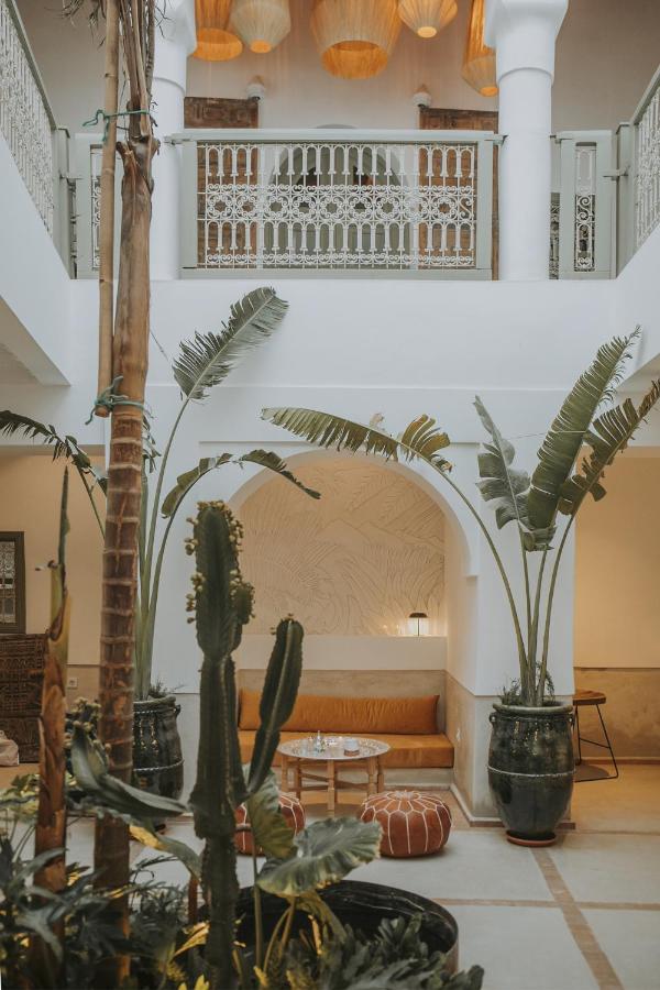 12 Charming Budget Riads in Marrakech 8 A stylish indoor courtyard in one of the budget riads in Marrakech features tall potted plants, a cactus, and palm trees, with cozy seating on yellow cushions under an arched wall. Ornate railings and warm hanging lights create an inviting atmosphere.