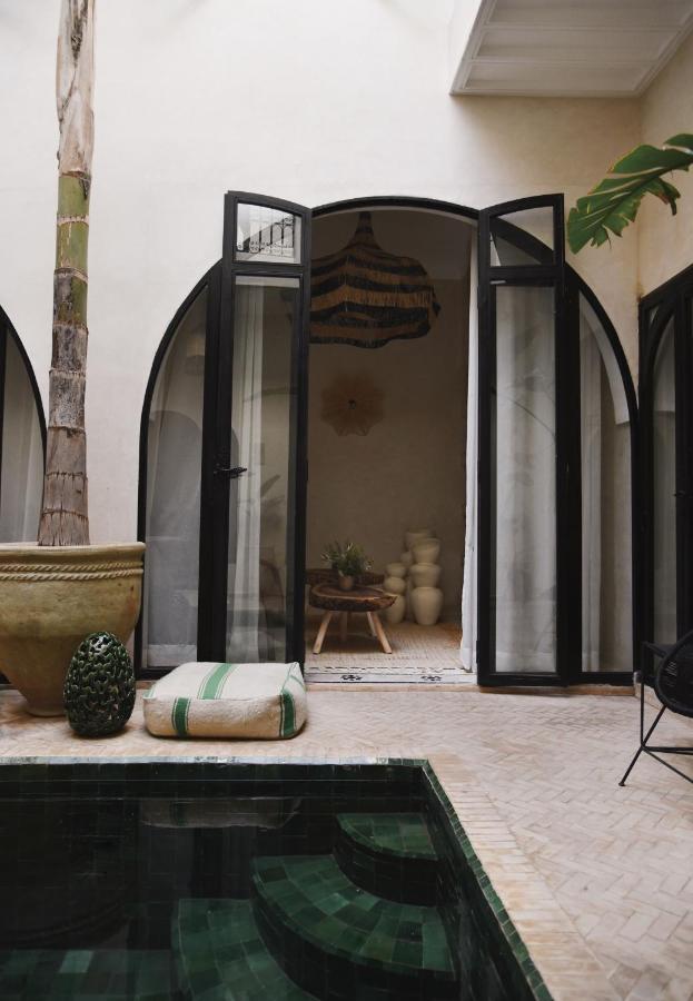 12 Charming Budget Riads in Marrakech 34 A serene courtyard in one of the charming budget riads in Marrakech features a small tiled pool, potted plants, a large green-and-white cushion, and arched black-framed glass doors opening to a cozy room with ceramics and a woven chandelier.