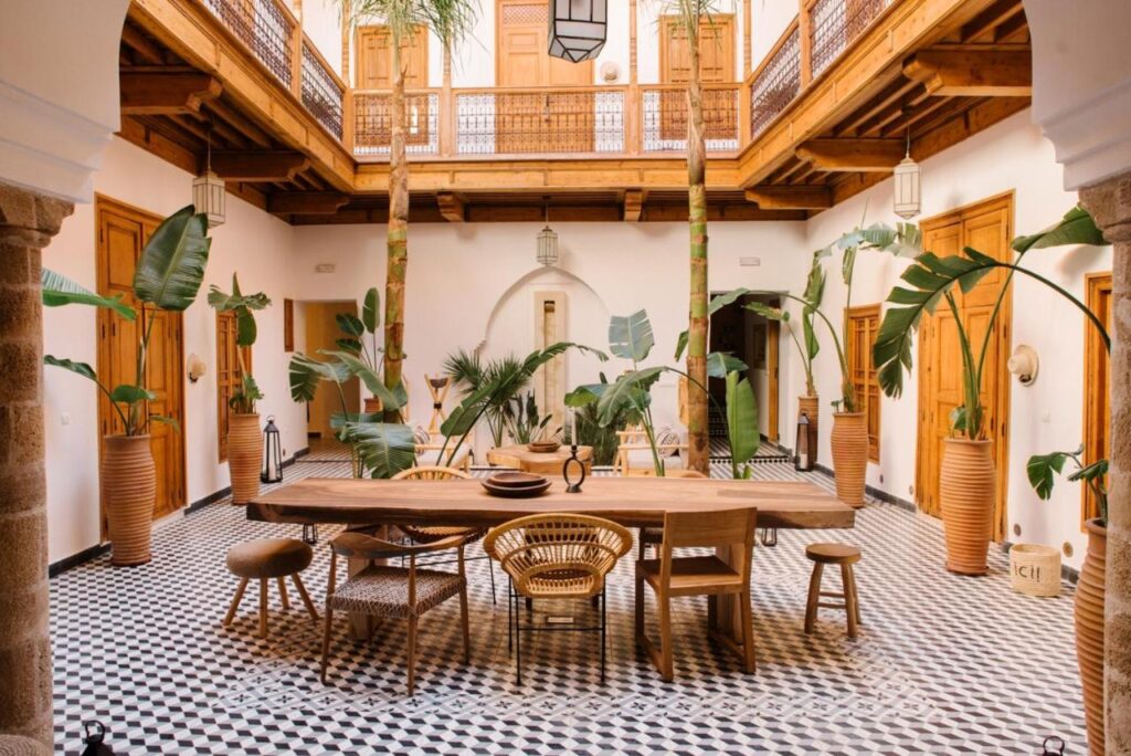 12 Charming Budget Riads in Marrakech 51 A spacious courtyard dining area in one of the budget riads in Marrakech features a long wooden table with mixed chairs, large potted plants, black-and-white patterned tile flooring, wooden balcony railings, and warm wooden doors.