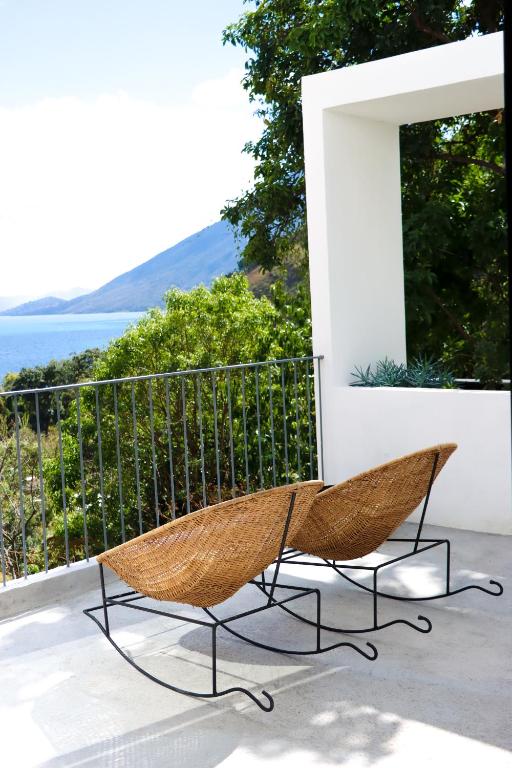 8 UNIQUE Places Where to Stay in Lake Atitlán 19 Two woven rocking chairs sit on a white patio with a metal railing, overlooking lush green trees and scenic Lake Atitlan views—an inviting spot for those searching where to stay in Lake Atitlan under a bright sky.