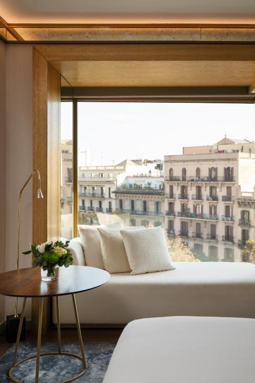 Top Barcelona Hotels with Rooftop Pool 47 A modern lounge area with a white chaise, pillows, a round wooden side table with a vase of flowers, and a large window offering a city view reminiscent of Barcelona hotels with rooftop pool vistas.