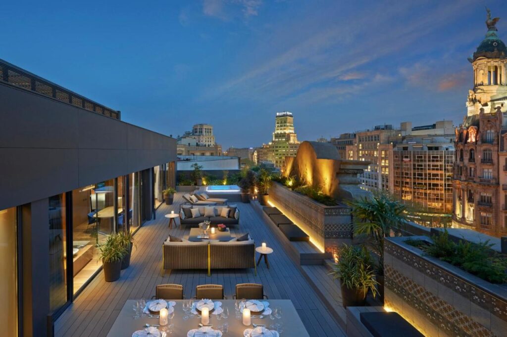 Top Barcelona Hotels with Rooftop Pool 21 A rooftop terrace at dusk features elegant outdoor seating, dining tables with white tablecloths, potted plants, and city buildings illuminated under a blue sky—an ambiance reminiscent of Barcelona hotels with rooftop pool views.