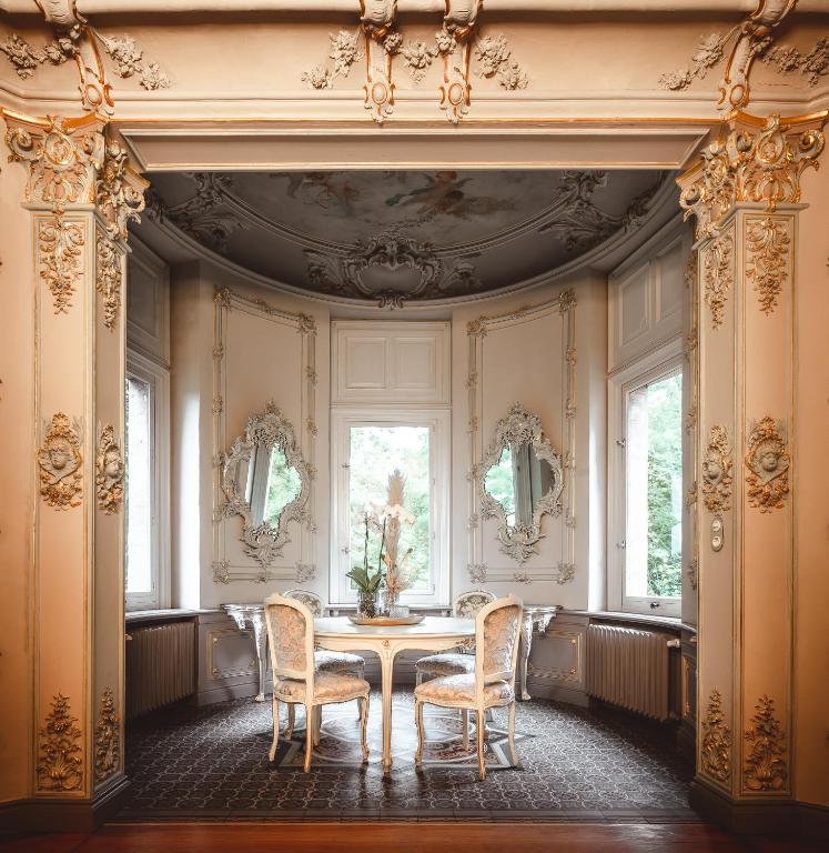 FAIRYTALE Castle Hotels in Bavaria 16 Ornate, vintage-style interior with detailed molding, two white chairs, and a small round table set by large, curved windows—reminiscent of the refined ambiance found in castle hotels in Bavaria. Elegant mirrors and floral decor enhance the luxurious atmosphere.