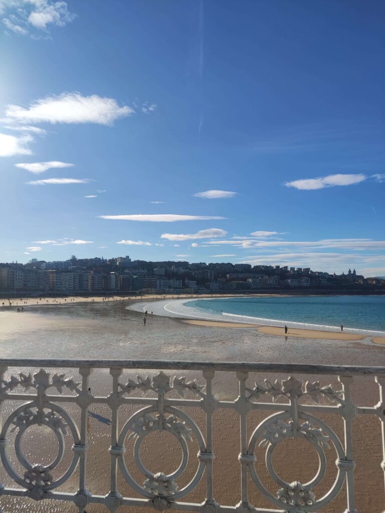 Best Boutique Hotels in San Sebastian 44 A view of a sandy beach with a few people walking along the shore, blue sea, and buildings—including boutique hotels in San Sebastian—in the background, seen over an ornate white railing under a clear, sunny sky with scattered clouds.