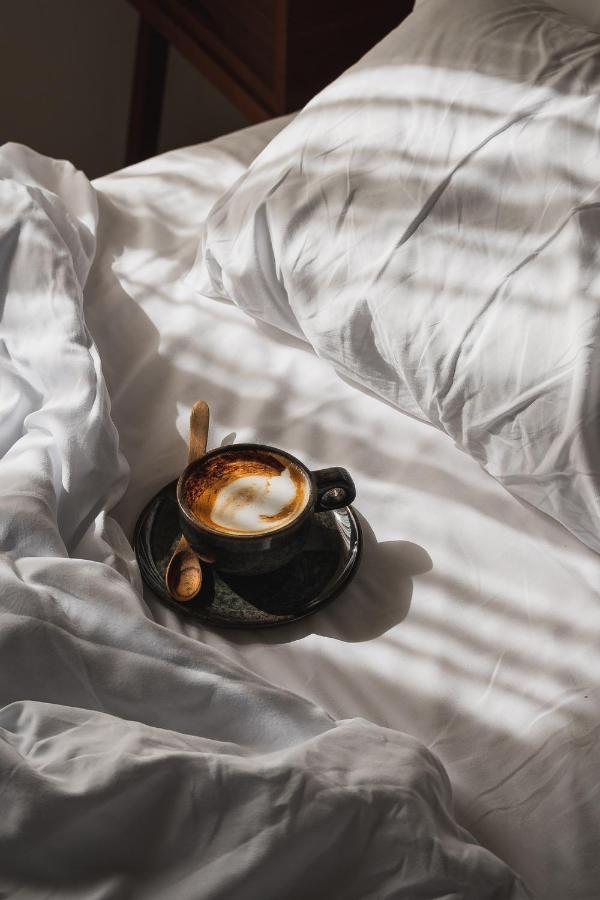 10 AUTHENTIC Finca Hotels in Ibiza 26 A cup of cappuccino with foam art sits on a saucer with a wooden spoon, placed on a rumpled white bed in one of the charming finca hotels in Ibiza, sunlight streaming through window blinds and casting shadow patterns on the sheets.