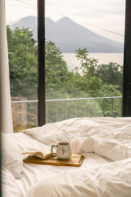 8 UNIQUE Places Where to Stay in Lake Atitlán 18 A cozy bed with white sheets and a wooden tray holding an open book and a mug sits by a large window—capturing the tranquil vibe many seek when searching where to stay in Lake Atitlan, with lush greenery, a calm lake, and mountains beyond.