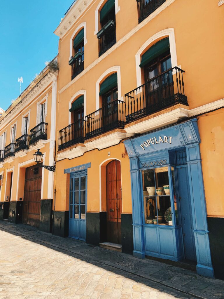 1 day in Seville Spain: My Favorite City 19 A vibrant yellow building with black balconies and arched green windows features a blue storefront labeled “POPULART,” displaying pottery inside—perfect to admire during 1 day in Seville, Spain as sunlight brightens the cobblestone street outside.