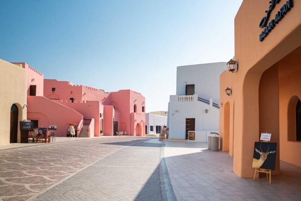 Where to Stay in Doha, Qatar: Transit, City & Beach Hotels 34 A sunlit, empty street lined with pastel-colored buildings, smooth arches, and balconies under a clear blue sky—this charming scene hints at where to stay in Doha, with art signs and outdoor furniture adding a welcoming touch.