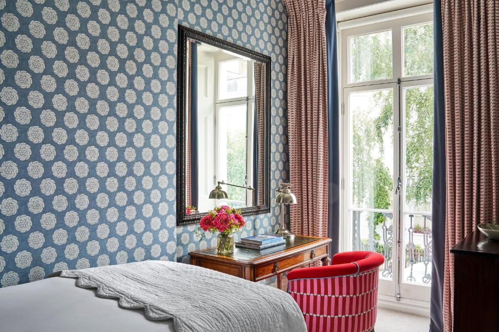 15 Best Boutique Hotels in London, UK 56 A cozy bedroom reminiscent of boutique hotels in London, with blue floral wallpaper, a large mirror above a wooden desk, a red striped chair, fresh flowers, and tall windows draped in pink curtains overlooking lush greenery.
