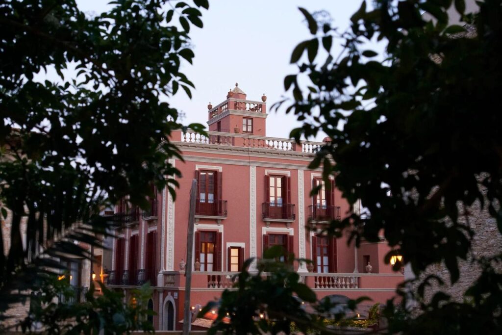 A pink, historic-style building with balconies and ornate detailing—like those found in boutique hotels in Costa Brava—is framed by green leafy branches in the foreground, viewed at dusk.