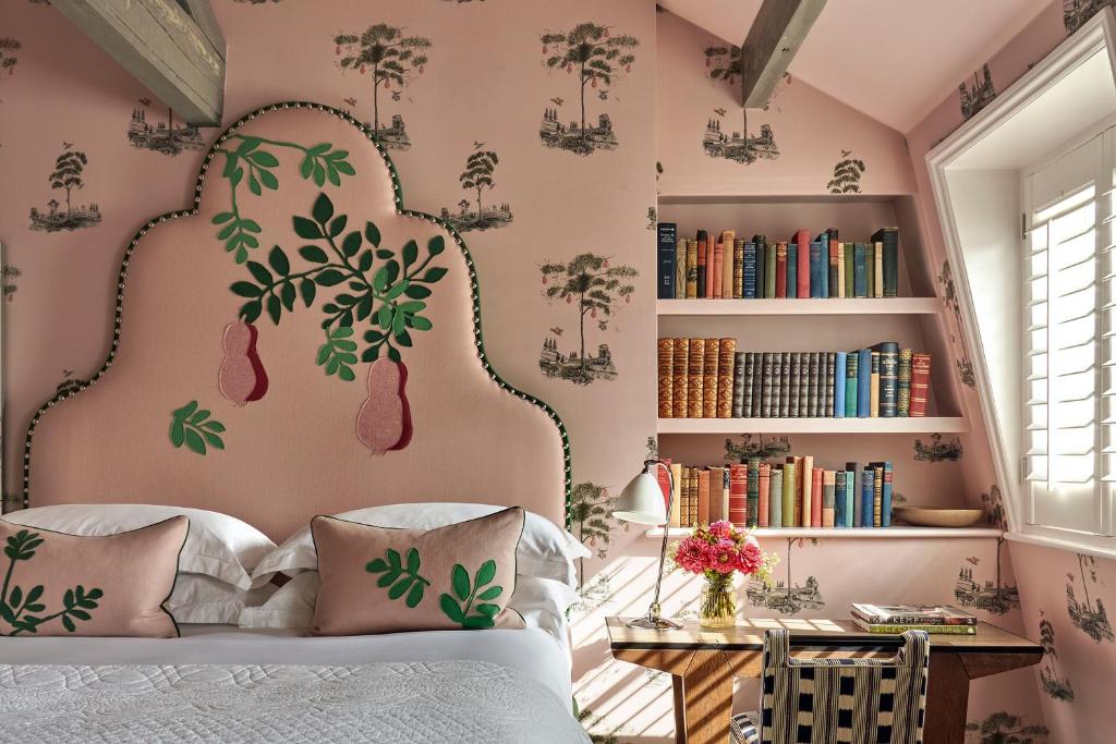 15 Best Boutique Hotels in London, UK 54 A cozy bedroom reminiscent of boutique hotels in London, with pink patterned wallpaper, a pink headboard adorned with green leaves and pears, crisp white bedding, a striped chair, a vase of flowers, and a bookshelf filled with colorful books.