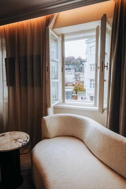 Unique Boutique Hotels in Salzburg 21 A cozy white curved sofa and a small round marble table sit beside an open window with sheer curtains, echoing the elegance found in boutique hotels in Salzburg and offering a view of white buildings and a hillside outside.