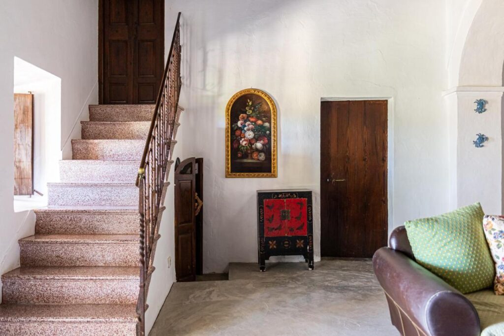 10 AUTHENTIC Finca Hotels in Ibiza 15 A cozy room in the style of finca hotels in Ibiza, featuring a brown staircase, wooden door, small floral painting above a decorative cabinet, and part of a brown sofa with green and yellow cushions. White walls and natural light brighten the space.
