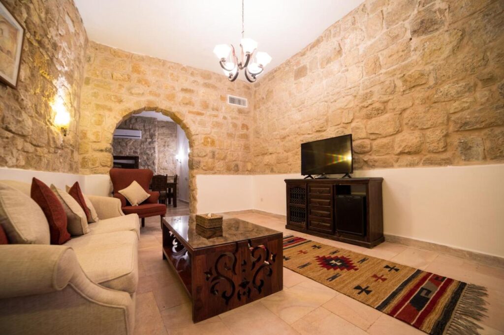 Authentic Hotels in Petra Jordan: Stay near the Ancient City 3 A cozy living room in the style of hotels in Petra, Jordan, featuring stone walls, a beige sofa with red and beige cushions, a glass-topped wooden coffee table, patterned rug, TV on a wooden stand, and an arched doorway to another room.