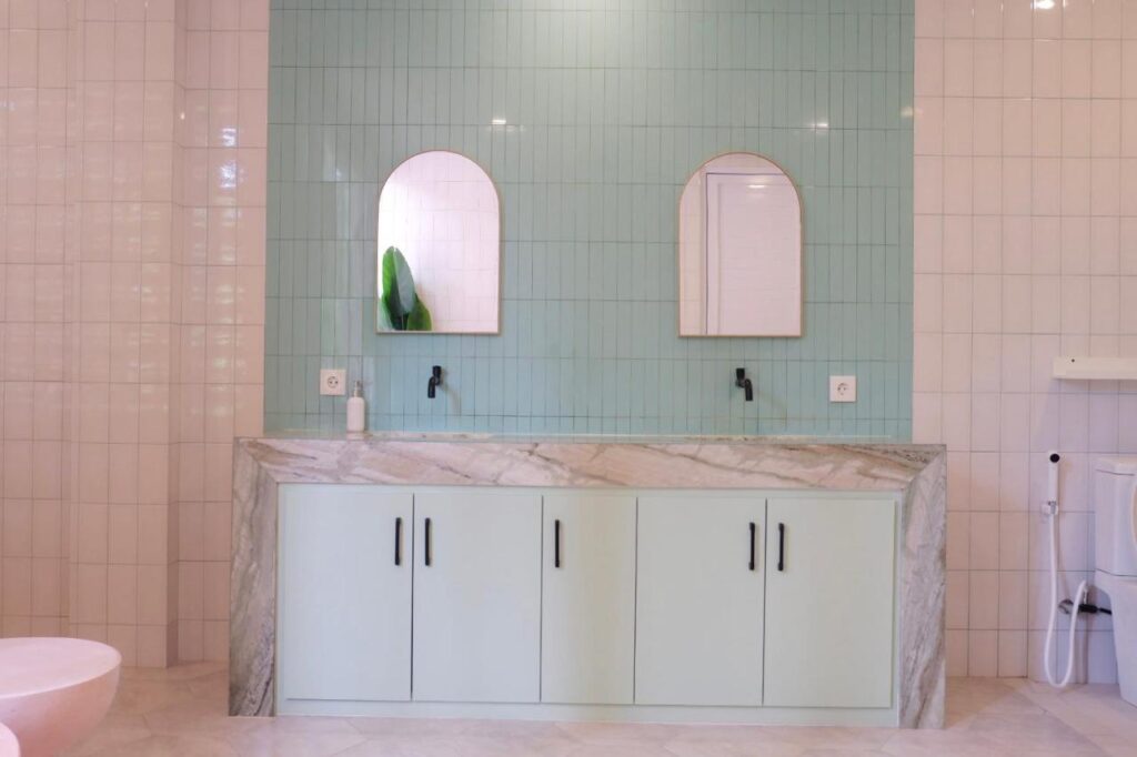 14 UNIQUE Boutique Hotels in Canggu, Bali 32 Modern bathroom with a double sink vanity featuring light green cabinets, black handles, and two arched mirrors—reminiscent of the stylish designs found in boutique hotels in Canggu. White and pale green tiled walls complement the minimalist black faucets.