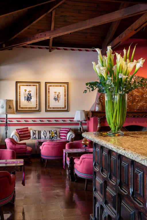 8 UNIQUE Places Where to Stay in Lake Atitlán 32 A cozy lounge—perfect for those seeking where to stay in Lake Atitlan—features pink armchairs, a granite counter topped with white calla lilies, framed artwork on cream walls, wooden beam ceiling, warm lighting, and vibrant decor accents.
