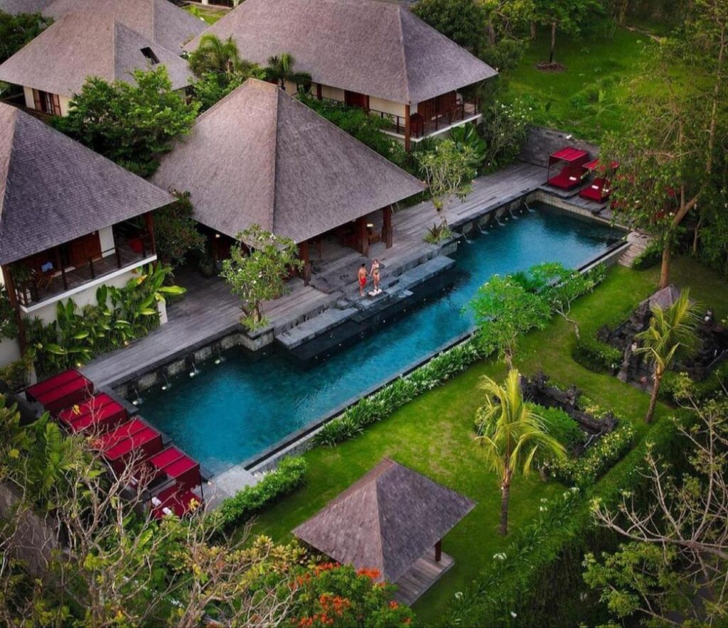 14 UNIQUE Boutique Hotels in Canggu, Bali 26 Aerial view of a tropical resort with villas, lush greenery, a long rectangular pool, red lounge chairs, and two people sitting by the poolside—perfect for those seeking boutique hotels in Canggu.