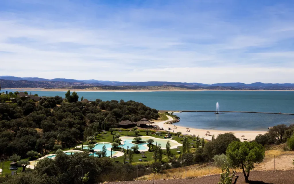25 Weekend Trips from Madrid Max. 2h Away 14 A scenic view of a lakeside resort, ideal for weekend trips from Madrid, featuring winding swimming pools, green lawns, trees, a sandy beach, and a distant pier with a water fountain set against rolling hills and a blue sky.