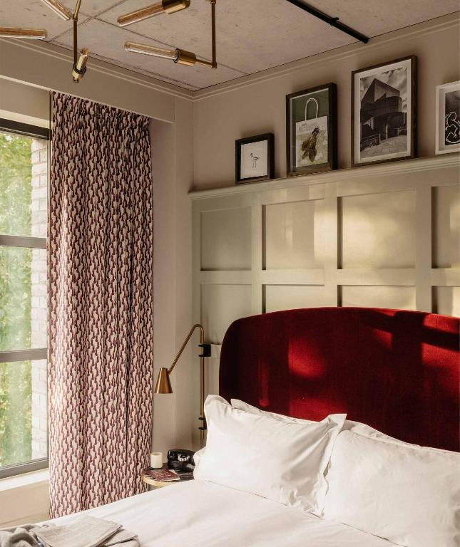 15 Best Boutique Hotels in London, UK 58 A cozy bedroom, reminiscent of boutique hotels in London, features a large white-paned window, patterned curtains, a deep red upholstered headboard, white bedding, a gold bedside lamp, and framed artwork above the bed as sunlight streams in.