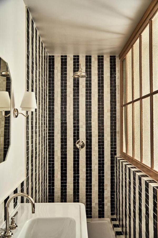 15 Best Boutique Hotels in London, UK 9 A small bathroom, reminiscent of the best boutique hotels London offers, features black and white vertical striped tiles, a white sink, wall-mounted faucet, round light fixture, and a window with wooden framing on the right side.