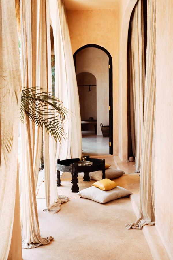 A cozy, sunlit patio in one of the best family villas in Bali features flowing beige curtains, a black low table with a tea set, floor cushions, a yellow pillow, an arched doorway to a softly lit interior, and a potted palm for greenery.