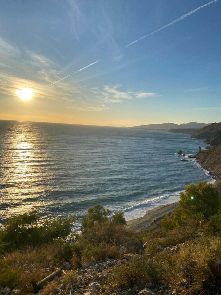 Adults-Only Hotels in Costa del Sol 41 Sunset over a calm ocean viewed from a cliff near adult only hotels in Costa del Sol, with gentle waves approaching a narrow beach below. Greenery and shrubs fill the foreground, and distant hills stand under a clear sky with streaks of clouds.
