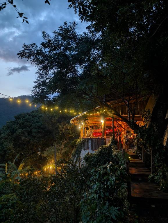 8 UNIQUE Places Where to Stay in Lake Atitlán 25 A cozy, warmly lit cabin nestled among dense trees at dusk—perfect for those seeking where to stay in Lake Atitlan—with string lights above a wooden walkway and a mountain visible beneath the darkening sky.