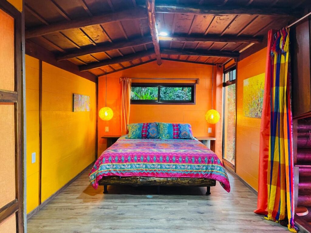 Unique Hotels in Costa Rica: Top Stays 19 A cozy bedroom with vibrant orange walls, a wooden ceiling, and a colorful bedspread. Like many unique hotels in Costa Rica, two glowing lamps brighten the space while a window above the bed lets in natural light from lush outside greenery.