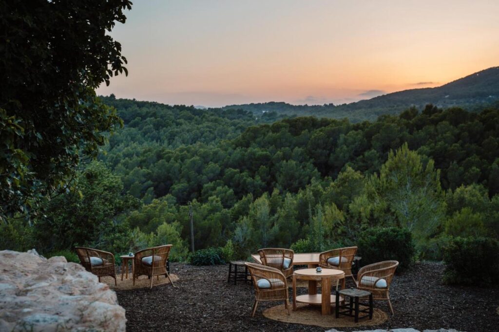 10 AUTHENTIC Finca Hotels in Ibiza 18 Wicker chairs and round tables are arranged outdoors on a terrace at one of the charming finca hotels in Ibiza, overlooking a lush, green forest with rolling hills at sunset. The sky is softly lit with orange and pink hues.