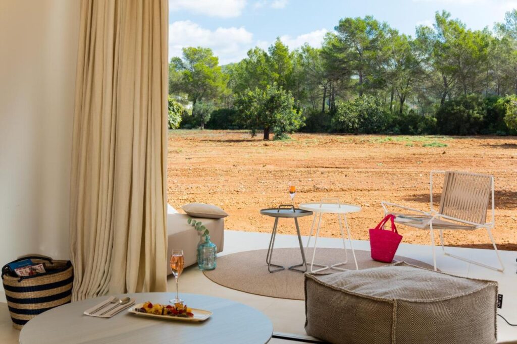 10 AUTHENTIC Finca Hotels in Ibiza 35 A cozy indoor seating area opens onto a patio with tables, a chair, and a pouf, offering views of the dry, earthy landscape typical of finca hotels in Ibiza. A pink bag and drinks are on the tables; a woven basket and food are inside.