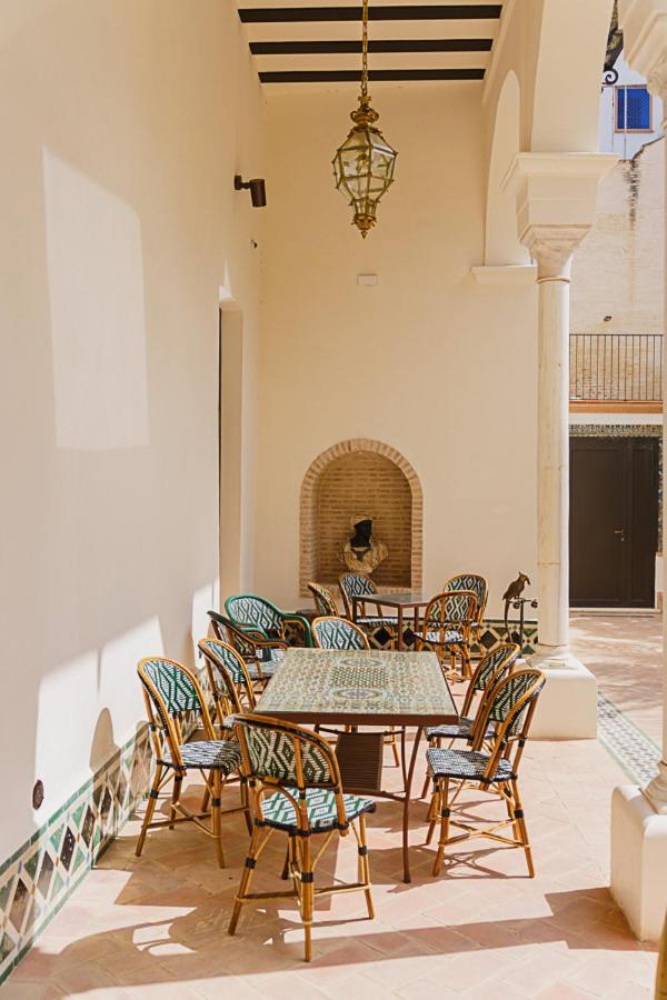 10 UNIQUE Hotels with Pool in Seville 37 Outdoor patio with a table and several patterned chairs, set against cream-colored walls and arches. Similar to many hotels with pool in Seville, a lantern hangs from the ceiling and a small wall fountain creates a tranquil backdrop. Sunlight casts gentle shadows on the floor.