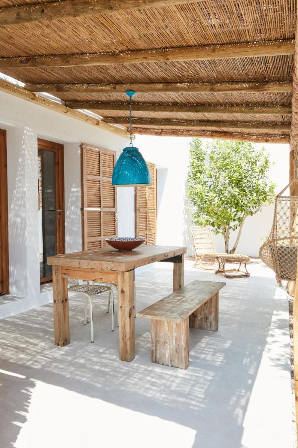 A rustic patio with a wooden table, matching bench, and stools sits under a pergola with bamboo slats—evoking the laid-back charm of boutique hotels in Formentera. A blue pendant light glows above, with wicker chairs and a sunlit tree in the background.