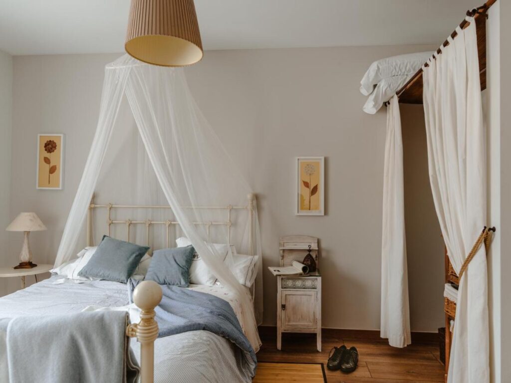 25 Weekend Trips from Madrid Max. 2h Away 75 Cozy bedroom with a white canopy bed, blue and white bedding, a small nightstand, framed floral prints, soft lighting, and a closet with curtains—perfect to unwind after weekend trips from Madrid. A pair of slippers rests on the wooden floor.