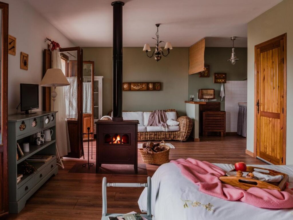 25 Weekend Trips from Madrid Max. 2h Away 74 Cozy rustic room with a wood stove, wicker sofa, and a bed with a pink blanket—perfect for relaxing weekend trips from Madrid. A tray with apples and drinks sits on the bed, while warm lighting and wooden accents create a welcoming atmosphere.