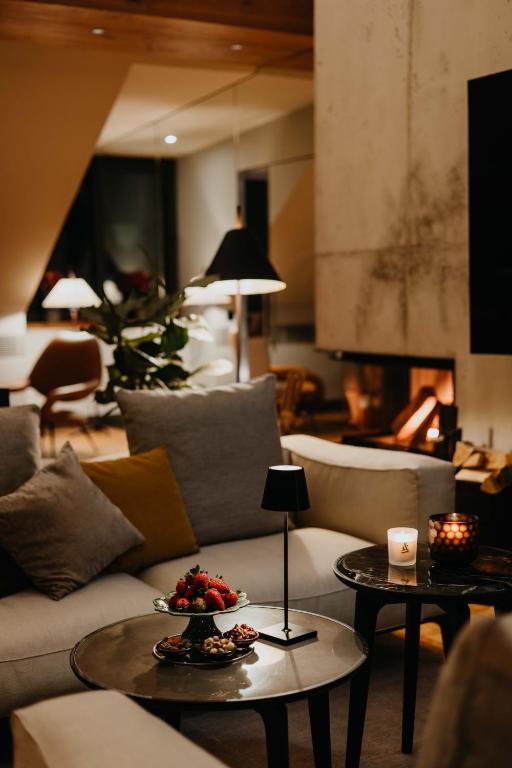 Authentic Boutique Hotels in Munich 21 A cozy living room with a modern design, inspired by boutique hotels in Munich, features a lit fireplace, gray sofas with cushions, round coffee tables holding a candle, a lamp, and fruit. Warm lighting and plants add to the inviting atmosphere.