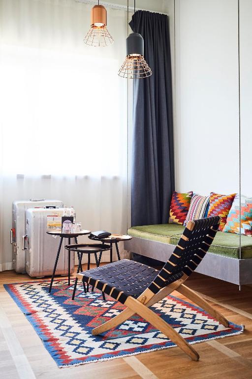 Authentic Boutique Hotels in Munich 7 Bright, modern living room reminiscent of boutique hotels in Munich, featuring a black woven lounge chair, patterned rug, small tables, green cushioned bench with colorful pillows, metal suitcase, and industrial-style pendant lights by sheer and dark curtains.