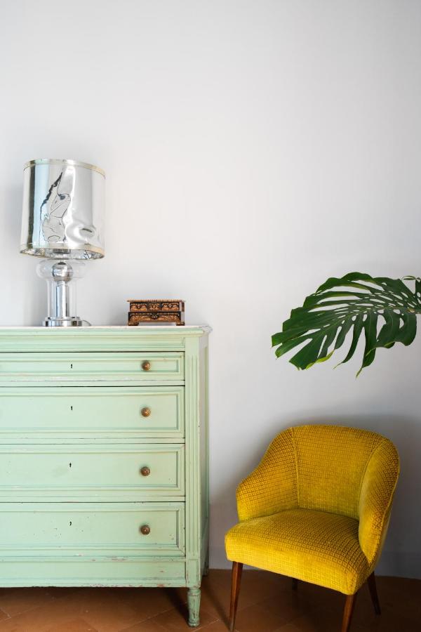 Boutique Hotels on the Amalfi Coast 7 A light green dresser with brass knobs holds a silver lamp and a small wooden box. Beside it is a yellow textured chair, echoing the vibrant charm of boutique hotels on the Amalfi Coast, while a large green leaf extends across the white wall.