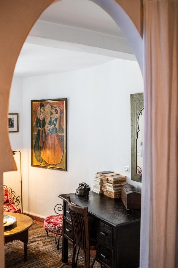 9 UNIQUE Riads in Tangier 15 A cozy room in the style of riads in Tangier, featuring a dark wooden desk, stacked books, vintage telephone, and wooden chair seen through an arched doorway. White walls display a painting and mirror, with patterned furniture completing the scene.